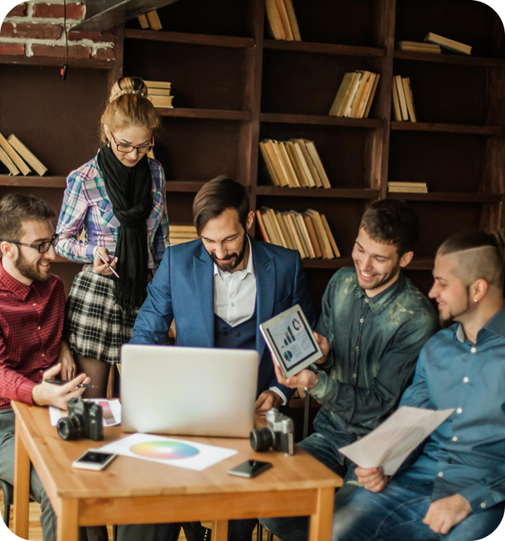 Group of five people sitting around a wooden table with laptops, tablets, and papers, in a cozy room with bookshelves